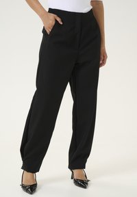 Pantalons noir cintrés avec une taille haute, des plis et des poches latérales, fabriqués dans un tissu lisse ; associés à des chaussures à talons noires.