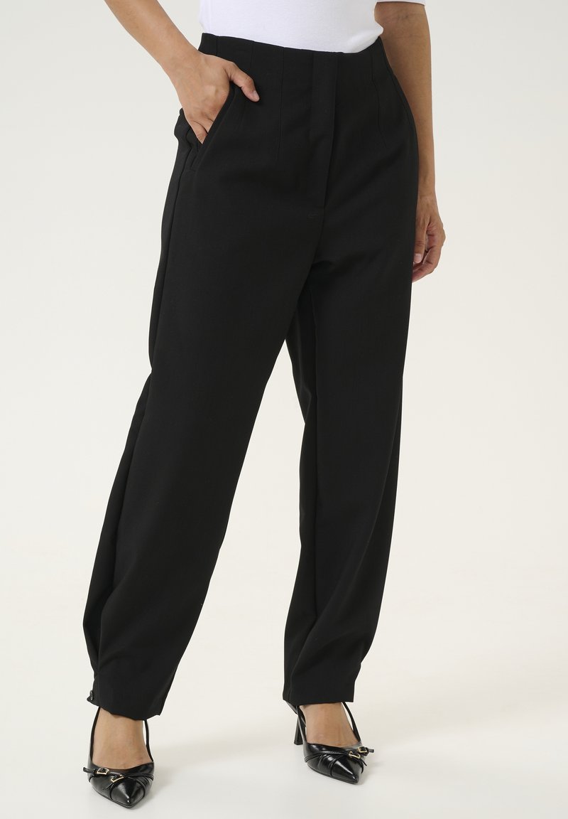 Pantalons noir cintrés avec une taille haute, des plis et des poches latérales, fabriqués dans un tissu lisse ; associés à des chaussures à talons noires.