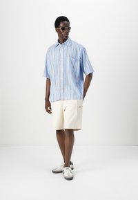Camisa de manga corta a rayas azules con líneas naranjas, combinada con pantalones cortos crema y zapatillas con detalles grises. Accesorios con gafas de sol claras.