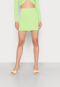 Top de malha canelada verde de mangas longas com design cropped e uma saia mini combinando com bainha em forma de ondas, acompanhados de mules amarelas de ponta aberta.