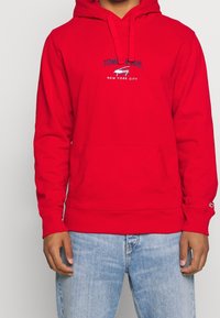 Felpa rossa con cappuccio e tasca frontale, caratterizzata da un logo ricamato blu che recita "TOMMY JEANS NEW YORK CITY." Polsini e orlo a coste.