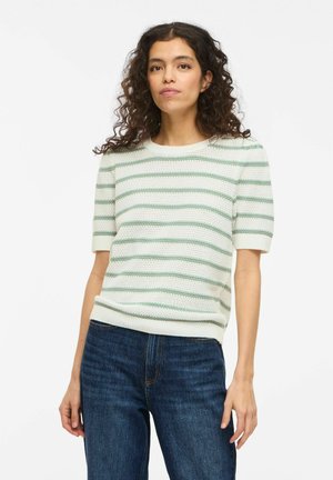 Femme aux cheveux bouclés portant un haut en maille blanc à manches courtes avec des rayures vertes et un jean bleu, debout devant un fond blanc.