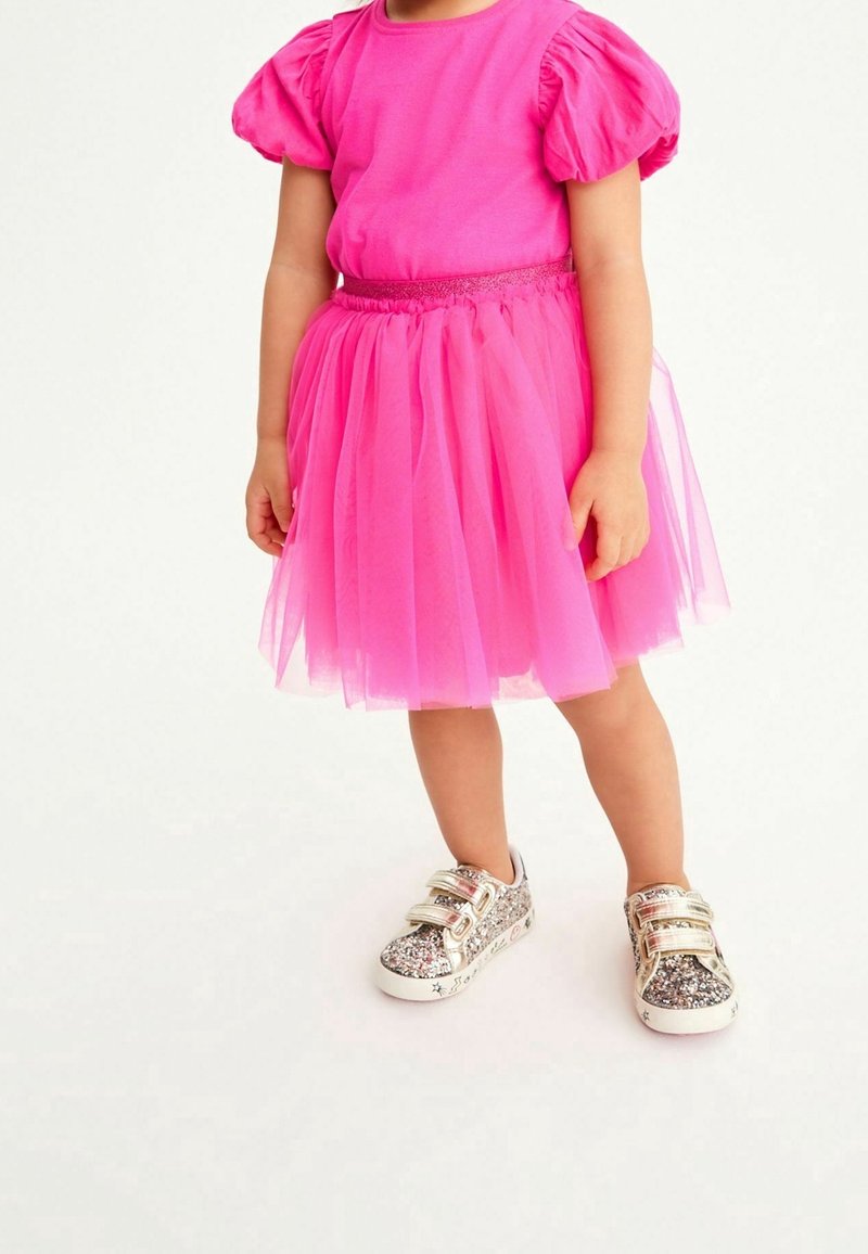 Enfant portant un haut à manches bouffantes rose vif et une jupe en tulle avec des baskets à scratch brillantes, debout sur un fond clair.