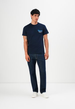 Jeune homme se tenant debout, portant un t-shirt imprimé bleu marine, un jeans bleu foncé et des baskets blanches sur un fond blanc uni.