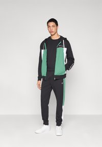 Μαύρο και πράσινο φούτερ με φερμουάρ, συνδυασμένο με μαύρη φόρμα που έχει πράσινες και λευκές πλαϊνές ρίγες. Λευκά αθλητικά παπούτσια ολοκληρώνουν το σύνολο.