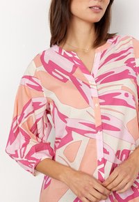 Blouse à motifs roses et pêche avec un devant boutonné, un col en V et des manches bouffantes. Fabriquée dans un tissu léger et texturé.
