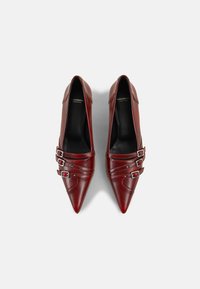 Zapatos de tacón alto de cuero rojo con diseño de punta afilada, que presentan múltiples hebillas decorativas y acentos plisados en los lados.