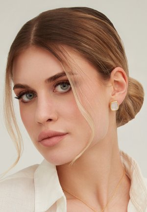 Femme aux cheveux châtain clair attachés en un chignon bas, portant des boucles d'oreilles en pierre blanche encadrées d'or et un délicat collier chaîne en or, vêtue d'une chemise blanche à col.