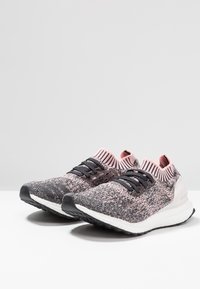 Pár růžových a černých pletených běžeckých bot s černými tkaničkami, bílými polstrovanými podrážkami a žebrovanými kotníkovými límci na bílém povrchu.