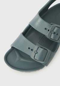 Grå plast-sandal med to justerbare stropper, teksturert overflate og en formet fotseng. Har spennebeslag for sikker passform.