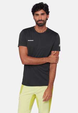 Uomo con capelli ricci scuri e barba, che indossa una maglietta nera Mammut e pantaloni giallo chiaro, in piedi con un braccio incrociato sull'altro.