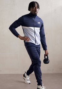 Marineblauer und weißer Sport-Trainingsanzug mit einer zipbaren Jacke und langen Hosen. Modell hält eine schwarze Kettlebell und trägt weiße Turnschuhe.