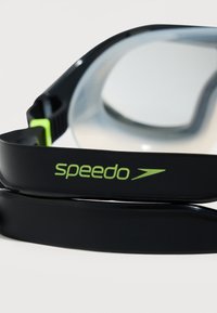 Μαύρα γυαλιά κολύμβησης με διαυγείς φακούς και πράσινο λογότυπο Speedo στο πλευρικό λουράκι, εμφανίζονται σε λευκό φόντο.