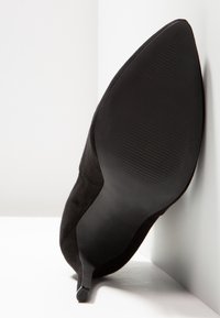 Černá semišová vysoká podpatek s špi�čatou špičkou a texturovanou podrážkou je nakloněna na bílém pozadí, což ukazuje na elegantní design.