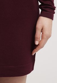 Robe pull bordeaux avec une texture lisse ; gros plan sur une main reposant sur l'ourlet, mettant en valeur le tissu et le design des poignets.