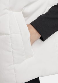 Biała kurtka puchowa z teksturowanym, pikowanym wzorem i boczną kieszenią. Pod spodem widoczna czarna koszulka z długim rękawem. Miękka, gładka tkanina.