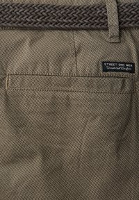 Section de pantalon en tissu beige à motif avec une poche arrière horizontale, une ceinture marron tressée et une étiquette noire "Street One Men".