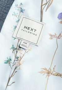 Balts audums ar gaišu ziedu rakstu un pelēku akcentu. Etiķetē rakstīts "NEXT TAILORING" un izmērs 40R; marķējums norāda "RAŽOTS BANGLADESHĀ".