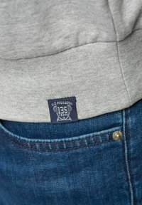 Grijze sweatshirt met een marineblauwe label met de tekst "U.S. POLO ASSN." en "135 JAAR." Blauwe denim jeans met een zichtbare messing knoopdetailing.
