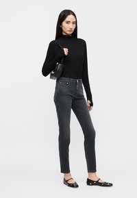 Femme portant un haut noir à manches longues, un jean taille haute gris foncé, des ballerines Mary Jane noires à clous, et portant un petit sac à bandoulière noir.