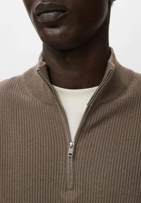 Suéter marrón de canalé con cuello alto con cremallera, que presenta una textura suave y se lleva sobre una camisa de color crema.