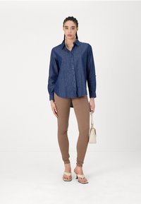 Chemise en jeans bleue à boutons avec manches longues, associée à un pantalon beige ajusté et des sandales à talons beiges. Tient un petit sac à main blanc.