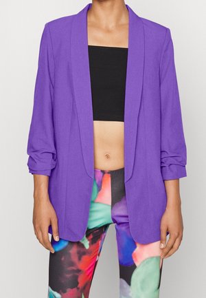 Femme portant un blazer violet sur un crop top noir et des leggings colorés à motif abstrait, debout devant un fond uni.