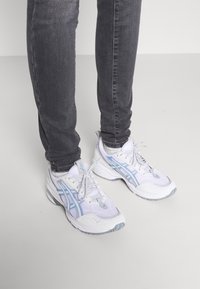 Vita och ljusblå löparskor med ovandel i mesh och syntetmaterial, med snörning och strukturerad sula, kombinerade med mörkgrå jeans.