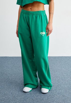 Osoba ubrana w luźne zielone spodnie dresowe Nike z białymi lamówkami, białe trampki oraz krótki zielony top, stojąca na szarym dywanie.