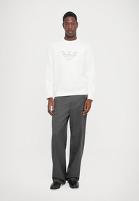 Hombre de pie que lleva una sudadera blanca con un pequeño logotipo estampado, pantalones anchos grises y zapatos negros, sobre un fondo blanco liso.