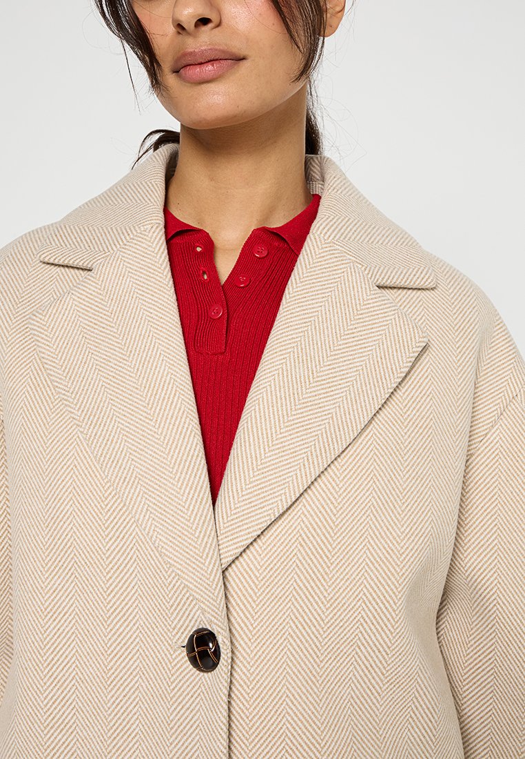 Beżowy płaszcz w jodełkę z dużym kołnierzem, zapinany na jeden czarny guzik, noszony z teksturowanym czerwonym polo pod spodem.