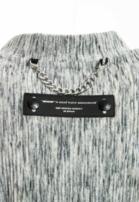 Suéter de rayas gris y blanco con un cuello alto, que presenta un detalle de cadena plateada y una etiqueta negra con texto y botones.