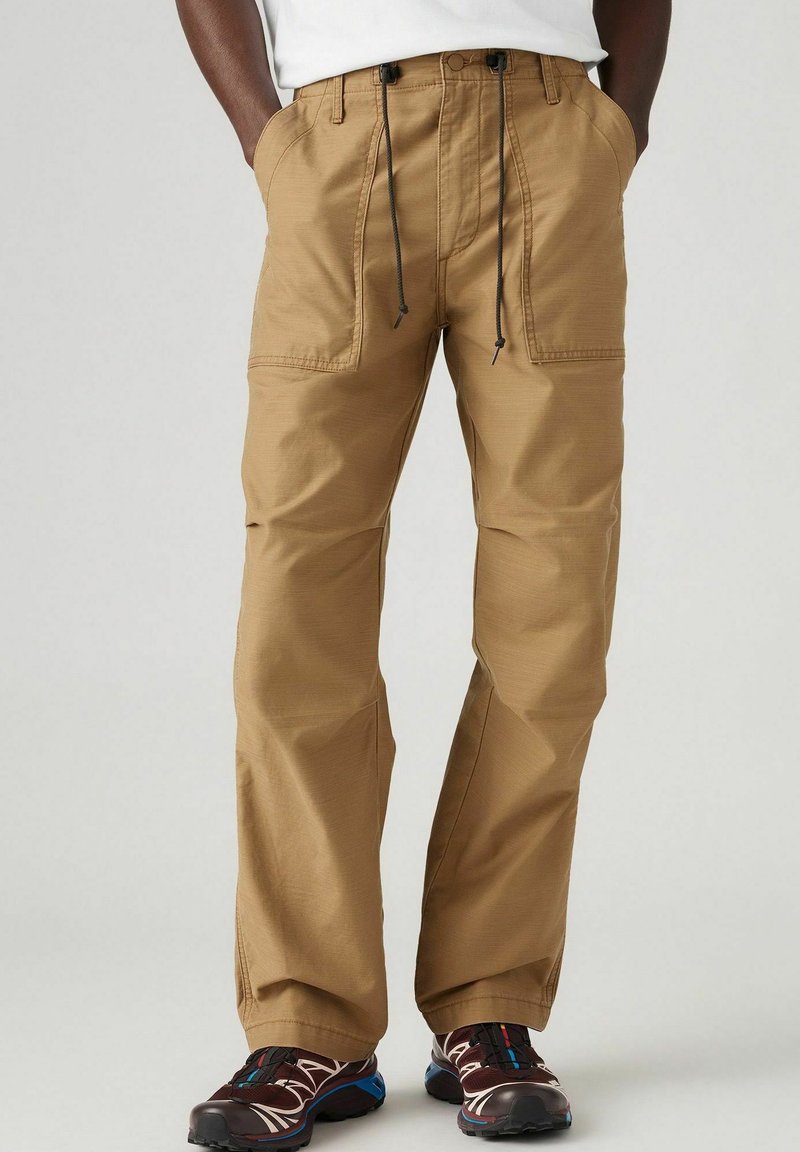 Pantalones cargo beige con un ajuste relajado, cintura con cordón y dos bolsillos frontales, fabricados en un tejido ligero adecuado para el uso casual.