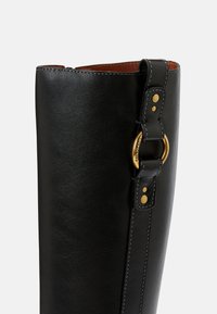 Stivale in pelle nera con texture liscia, caratterizzato da un'accento di anello dorato e una cintura dettagliata fissa con due bottoni dorati.