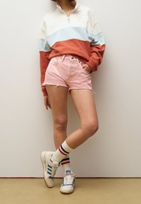 Persona vistiendo un sudadera de media cremallera en bloques de color, pantalones cortos de mezclilla rosa, calcetines de rayas y zapatillas blancas con detalles azules, de pie contra una pared.