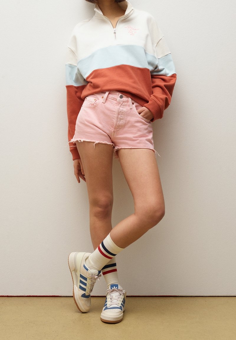 Persona vistiendo un sudadera de media cremallera en bloques de color, pantalones cortos de mezclilla rosa, calcetines de rayas y zapatillas blancas con detalles azules, de pie contra una pared.