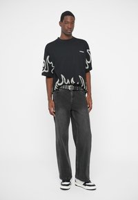 Mees, kes kannab musta leekide printimisega T-särki, musta metallist vööga, laiu musti teksaseid ja musta-valgeid sneidereid, seisab valge tausta ees.