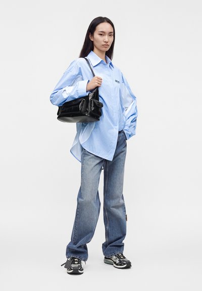 Jeune femme portant une chemise bleu clair ample, un jean large, des baskets, tenant un sac à main en cuir noir sur l'épaule, debout devant un fond blanc.