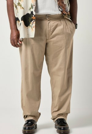 Hombre con pantalones beige plisados, zapatos de cuero negros, una camiseta blanca acanalada y una camisa estampada con diseños de tigres y bambú.