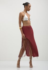 Jupe rouge avec des franges sur les côtés, associée à un haut de bikini blanc en forme de triangle. L'ensemble présente une fente haute, mettant en valeur des bretelles fines et une texture lisse.