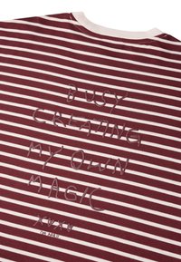 Juostuotas bordo marškinėliai su baltomis juostelėmis, su iškilniu tekstu: "UŽIMTA KURIANT SAVO MAGIJĄ" ir maža grafika apačioje.