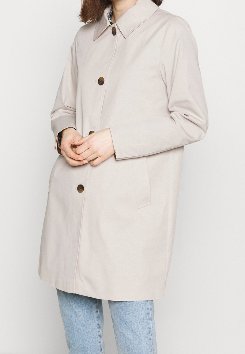 Beige regnrock med krage, framknappar och sidofickor. Tillverkad av ett slätt material med avslappnad passform.