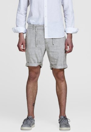 Homme portant un short à rayures gris clair retroussé, une chemise blanche à manches longues avec les poignets retroussés, et des baskets grises, debout devant un fond uni.
