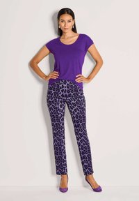 Vrouw staat met handen op de heupen in een paarse top, paarse luipaardprint broek en paarse hakken tegen een lichte achtergrond.