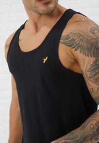 Top in cotone nero senza maniche con scollatura rotonda e un piccolo logo dorato di un uccello sul petto. Il tessuto è liscio con una leggera elasticità.