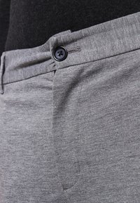 Grå tygbyxor med en strukturerad yta, med svart knäppning och fickor på sidorna. Midjebandets design är slät med lätt rynkning.