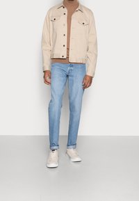 Giacca di jeans beige con bottoni scuri, jeans azzurri chiari e sneakers color crema. L'outfit presenta un design semplice e una vestibilità rilassata.