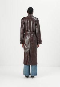 Brauner Kunstleder-Trenchcoat mit Taillengürtel, langen Ärmeln und Schulterepauletten, kombiniert mit blauen Jeans im weit geschnittenen Stil und schwarzen High Heels.