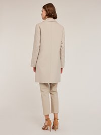 Beige Mantel mit Reverskragen, mittellang und geradem Schnitt. Kombiniert mit hellbeigen Hosen und braunen Riemchen-Heels.