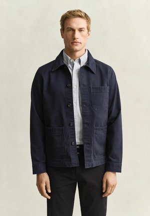 Blonder Mann trägt eine dunkelblaue, geknöpfte Jacke über einem hellgrau gestreiften Hemd und steht mit neutralem Gesichtsausdruck vor einem einfarbigen Hintergrund.